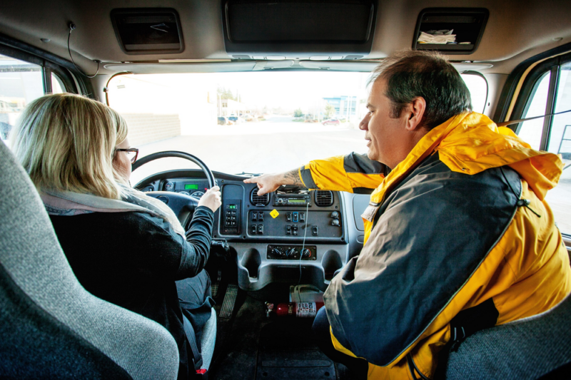 A man in a yellow jacket points at the dashboard of a truck while talking to a woman sitting in the driver’s seat. The woman is looking at where he is pointing. The interior of the truck cab is visible, with the steering wheel and various controls in view. A man in a yellow jacket points at the dashboard of a truck while talking to a woman sitting in the driver's seat. The woman is looking at where he is pointing. The interior of the truck cab is visible, with the steering wheel and various controls in view.
