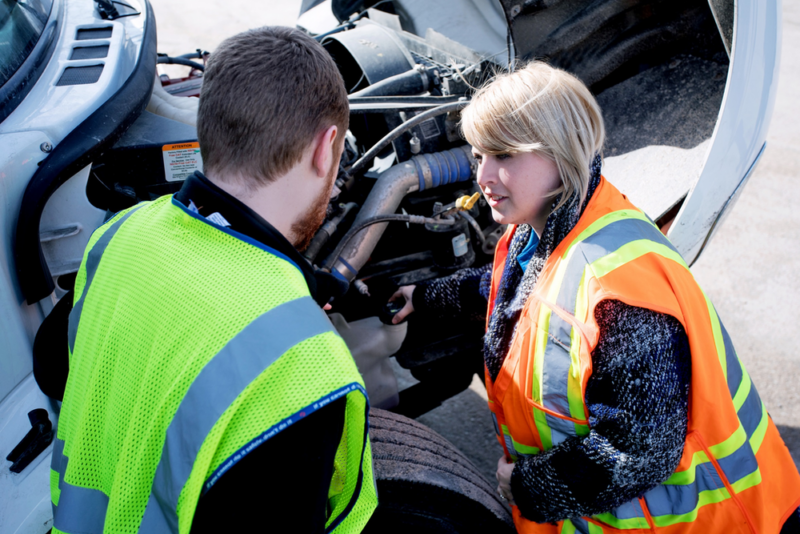 Two people wearing high-visibility vests inspect the engine of a large truck. The individual on the left, wearing a yellow vest, appears to be explaining something, while the person on the right, in an orange vest, listens attentively. The truck hood is open. Two people wearing high-visibility vests inspect the engine of a large truck. The individual on the left, wearing a yellow vest, appears to be explaining something, while the person on the right, in an orange vest, listens attentively. The truck hood is open.