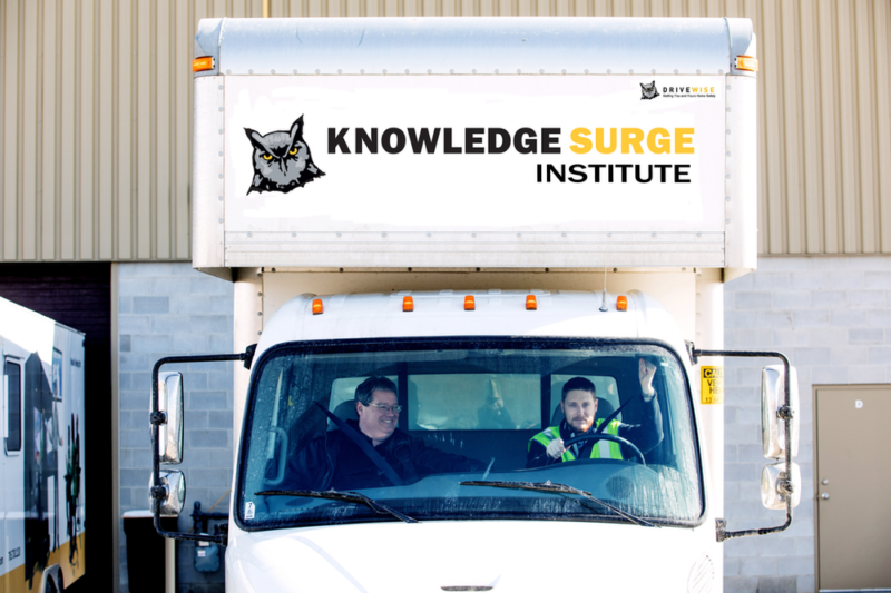 A truck with the “Knowledge Surge Institute” logo on its front box is parked. Two people are seated inside the truck, smiling. The truck is in front of a building with a beige exterior and a brown garage door. Another truck is visible in the background on the left. A truck with the "Knowledge Surge Institute" logo on its front box is parked. Two people are seated inside the truck, smiling. The truck is in front of a building with a beige exterior and a brown garage door. Another truck is visible in the background on the left.