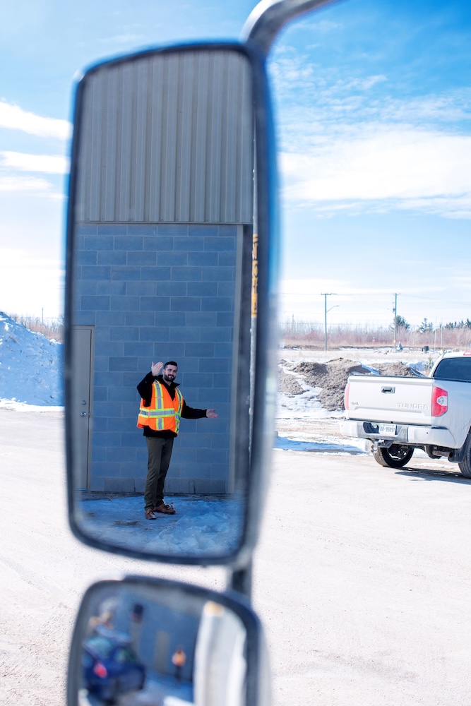 A man wearing a bright orange safety vest and winter clothing is reflected in the rearview mirrors of a vehicle. He is standing outdoors on a snowy day, in front of a blue building and beside a white truck. He is waving and smiling.