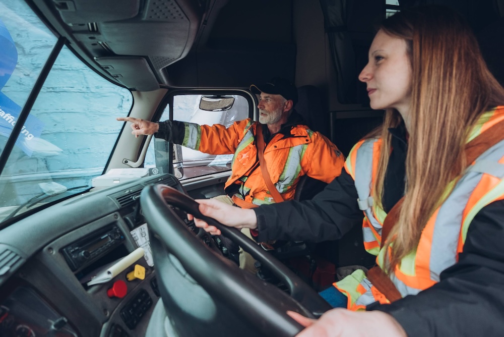 A man and a woman, both wearing orange reflective safety gear, are inside a vehicle. The man, seated in the passenger seat, is pointing out something through the windshield, while the woman is seated in the driver’s seat, holding the steering wheel.