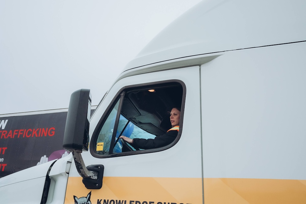 A person sits in the driver's seat of a large white truck, looking out of the window. Part of the truck's yellow stripe and black text are visible. In the background, there is a billboard with a message about stopping human trafficking.