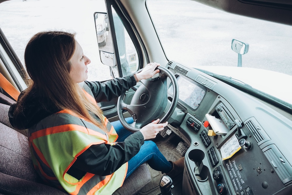 A person wearing a reflective safety vest sits in the driver's seat of a large vehicle, holding the steering wheel. The dashboard is visible, showing standard vehicle controls and instruments. The person appears to be focused on driving.