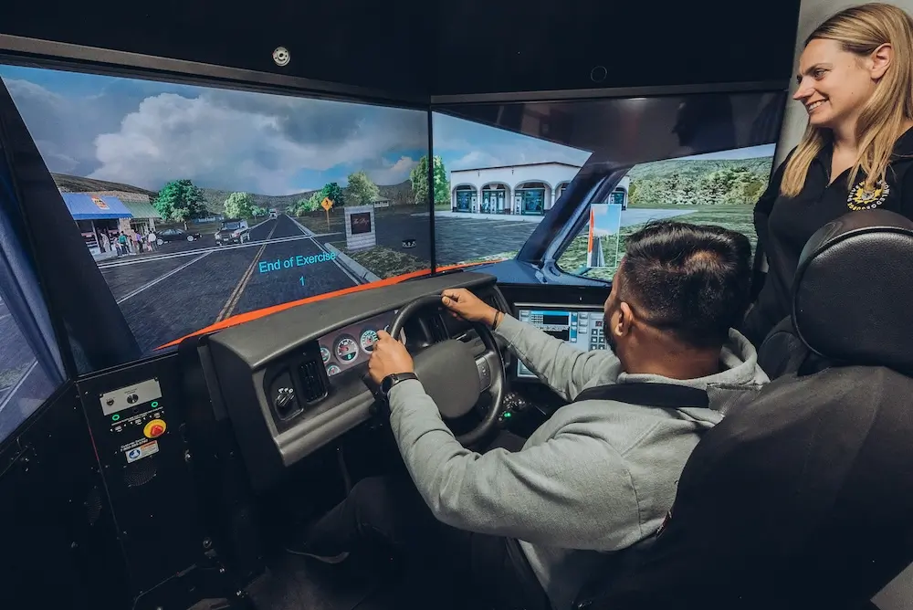 A person is seated in a driving simulator, holding the steering wheel. A large screen displays a virtual road scene. Another person stands beside the simulator, observing. The setting appears educational or training-focused.