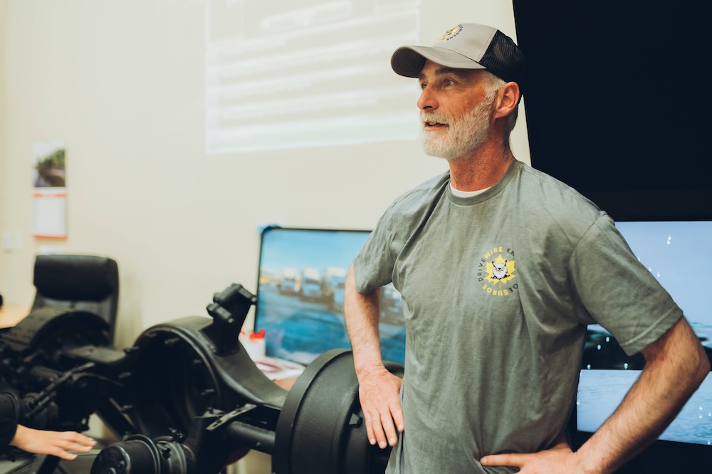 A man wearing a grey t-shirt and a cap stands with his hands on his hips, looking to the side. Behind him is a large screen displaying images and text, and to his left is a mechanical apparatus. An office chair and desk are partially visible.