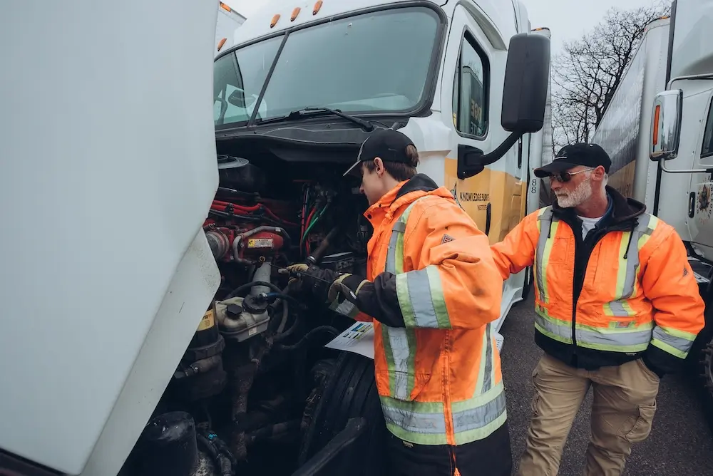 Knowledgeable-teacher-trucking-KnowledgeSurge Two people in reflective orange jackets examine the open engine of a large white truck. One person is working on the engine while the other observes. Several trucks are parked nearby on a rainy day.