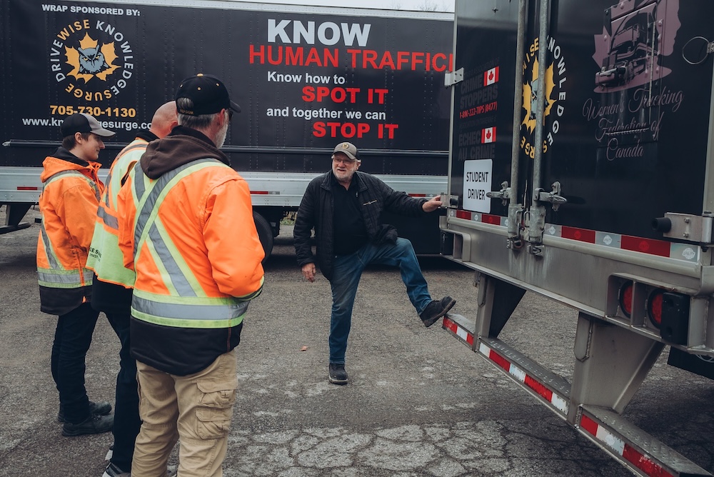 A man stands with one foot on the back of a truck, talking to three other men wearing high-visibility jackets. The truck has signs promoting awareness about human trafficking and encouraging people to learn how to spot and stop it.