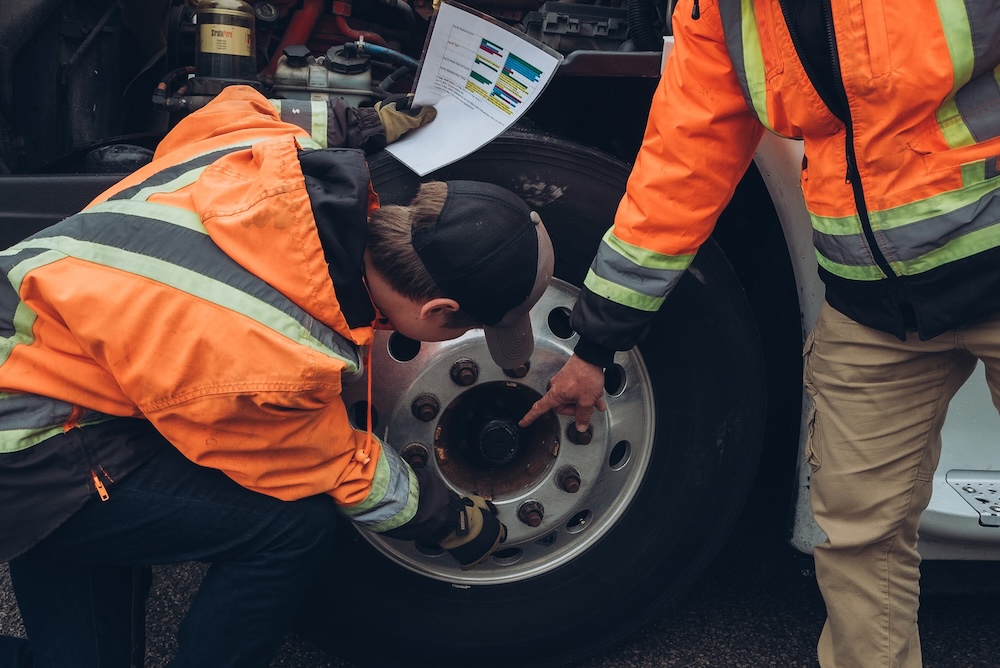 Two people wearing orange high-visibility jackets are inspecting a large vehicle tire. One person is pointing at a part of the tire while holding a paper with colorful charts. The other is looking closely at the tire's components with tools.