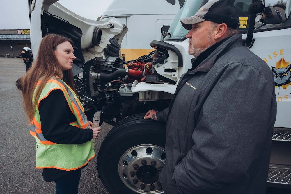 A woman in a fluorescent safety vest and a man in a jacket and cap stand beside a truck with its hood open, examining the engine. They appear to be discussing something related to the truck’s maintenance or inspection. A woman in a fluorescent safety vest and a man in a jacket and cap stand beside a truck with its hood open, examining the engine. They appear to be discussing something related to the truck's maintenance or inspection.