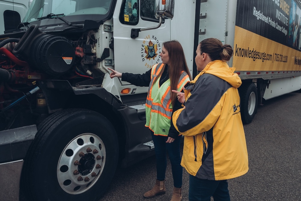 Two women in safety vests inspect the engine of a large truck. One is pointing to a part inside the engine compartment while discussing it with the other. The truck has a yellow trailer with writing on the side. They are standing on a paved surface.