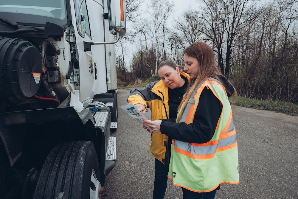 Two women stand by a large truck on a road surrounded by trees without leaves. They are examining a document together. One woman is wearing a yellow jacket, and the other is wearing a reflective safety vest over a black jacket.