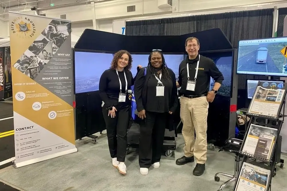 Three people stand and pose for a photo in front of a driving simulator booth at an event. The booth features informational banners, a large screen setup, and a monitor displaying a driving scene.