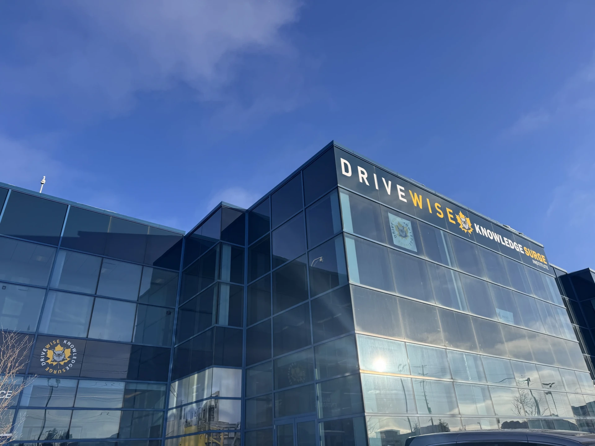 A modern glass office building with “DRIVEWISE KNOWLEDGE SURGE” signage on the upper corner, reflecting a clear blue sky. The building features large windows and a yellow logo near the entrance.