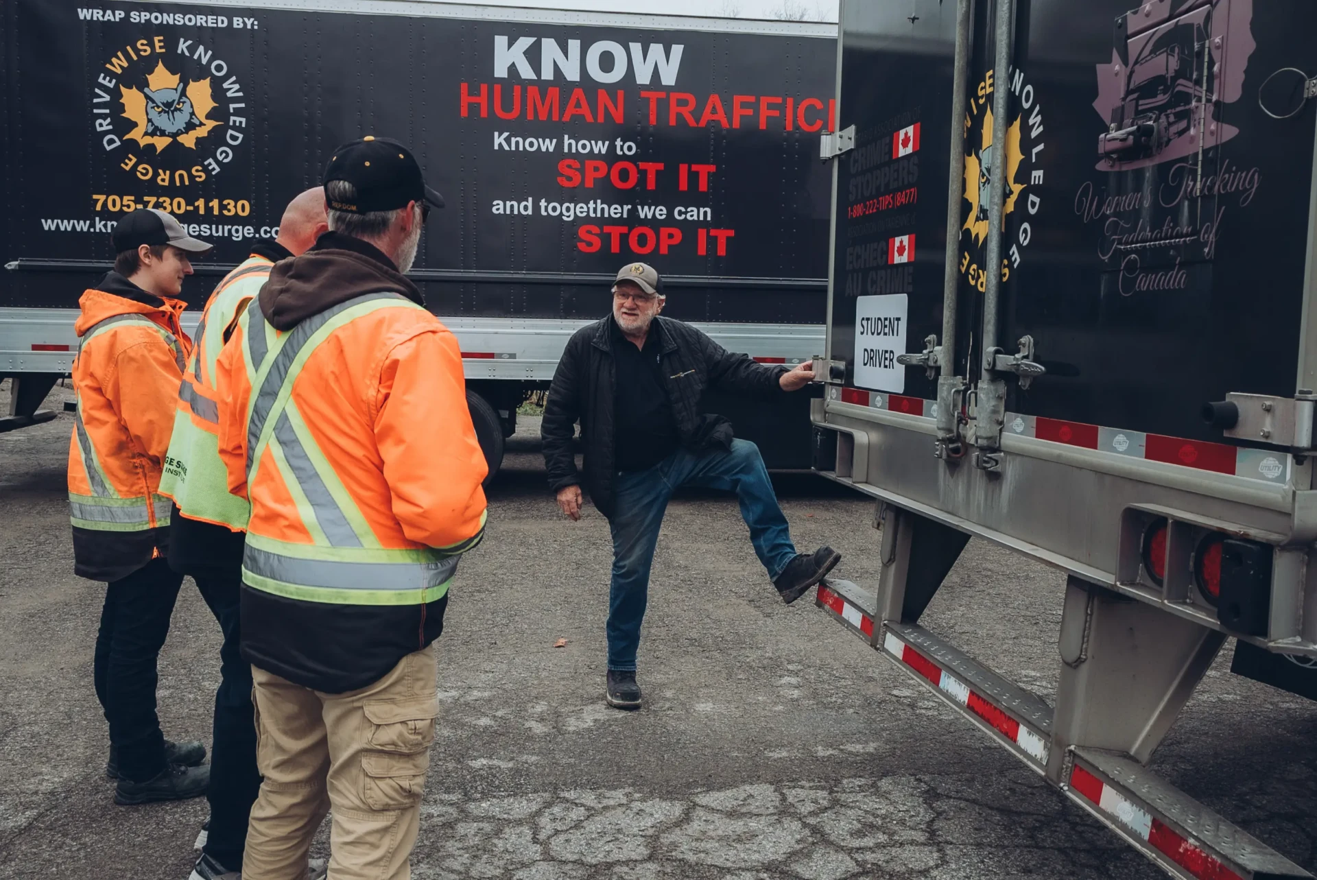 KnowledgeSurge Trucking School Three people in safety vests watch a man demonstrating something on a truck trailer. A large sign about spotting and stopping human trafficking is visible in the background.