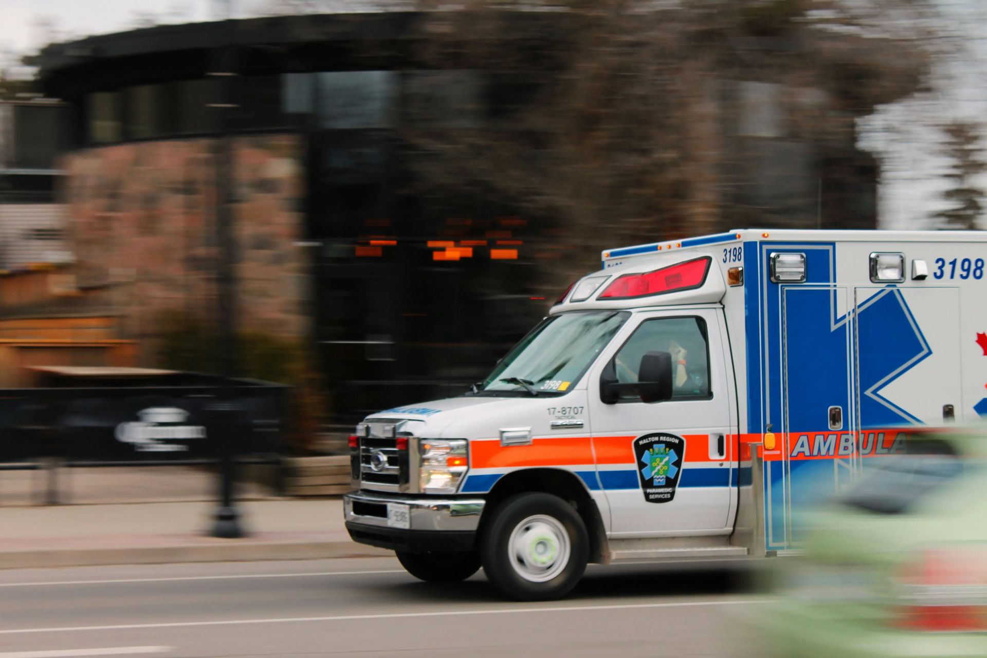 Ambulance with blue, orange, and white markings drives quickly on a city street. The background appears blurred, indicating motion, and a green vehicle is partially visible in the foreground.